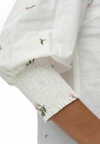 Indumento in cotone bianco con maniche a sbuffo e polsini arricciati ed elasticizzati. Presenta delicate ricami floreali rosa e foglie verdi in tutto il capo.