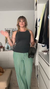 Sandali neri tenuti in una mano; indossa una canotta scura e pantaloni a righe verdi. Sullo sfondo si vedono scaffali con abbigliamento e decorazioni.