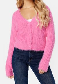 Pink, fluffy cardigan with a ribbed texture, v-neckline, and five buttons. Long sleeves feature fuzzy cuffs. Worn over dark denim pants.