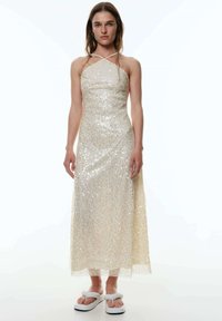 Paillettenverziertes, cremefarbenes, bodenlanges Kleid mit Halterneck und transparenter Saumkante. Kombiniert mit weißen Flip-Flops.