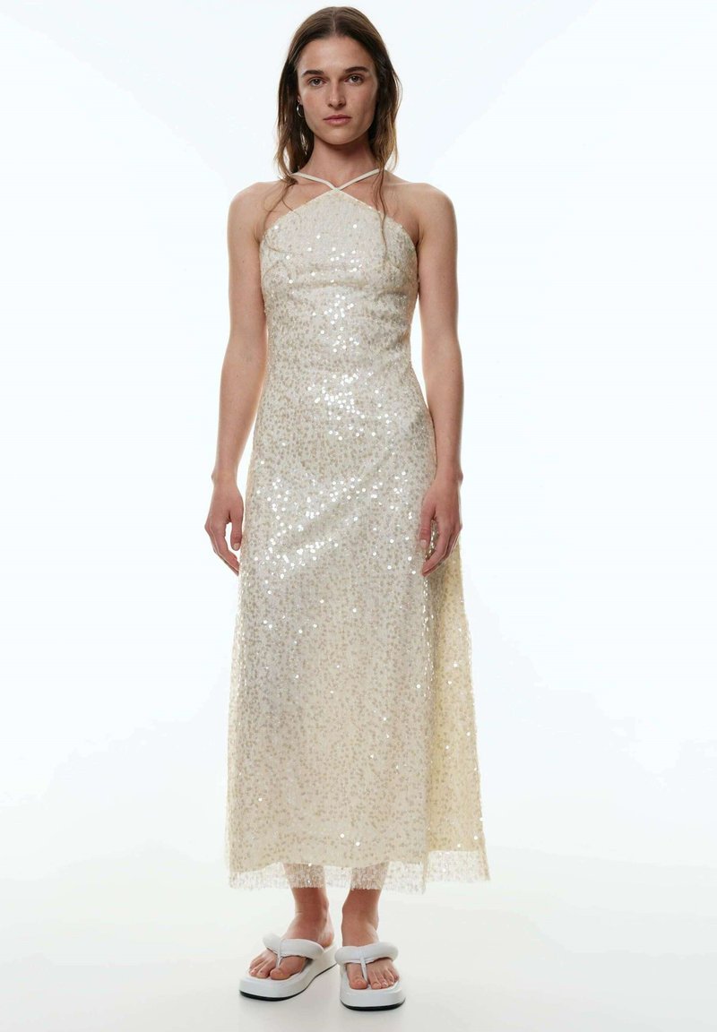 Paillettenverziertes, cremefarbenes, bodenlanges Kleid mit Halterneck und transparenter Saumkante. Kombiniert mit weißen Flip-Flops.