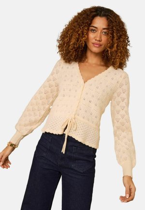 Žena s kučeravými vlasmi, oblečená v krémovom pletenom kardigáne s balónovými rukávmi a tmavých džínsoch s vysokým pásom, na neutrálnom pozadí.