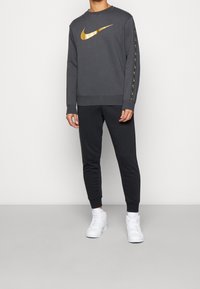 Grå sweatshirt med guld Nike-swoosh, med svarta byxor och vita sneakers. Sweatshirten har långa ärmar och ribbade muddar.