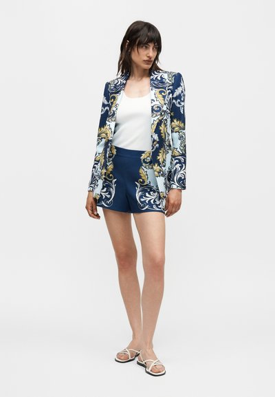 Γυναίκα με ναυτικό μπλέ σακάκι και τόνικ σορτς με χρυσά και λευκά μπαρόκ μοτίβα, λευκή αμάνικη μπλούζα και λευκά σανδάλια με λουράκια, όρθια.