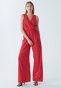 Rød heldragt med dyb V-udskæring, viddeben, justerbart taljebånd og glat tekstur. Kombineret med sorte højhælede sandaler.