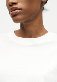 Κοντινή λήψη του κάτω μέρους του προσώπου και του λαιμού ενός ατόμου που φοράει ένα λευκό πουκάμισο με μικρές υφασμάτινες κουκκίδες.