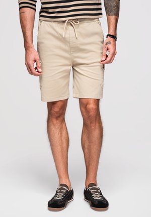 Hombre que lleva shorts beige con cordón, una camiseta de manga larga a rayas y zapatillas negras casuales con cordones blancos, de pie frente a un fondo liso.