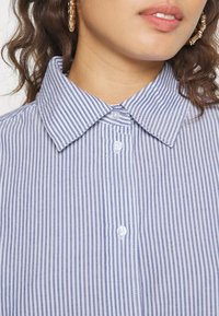 Sininen ja valkoinen raidallinen kauluspaita, jossa on napit; sis�ältää kevyen, kudotun kankaan ja klassisen leikkauksen. Korostuksia ovat muotoiltu kaulus.