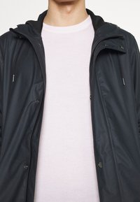 Mörk marinblå lättviktsjacka med huva, med dragkedja och tryckknappar, lager på lager över en ljusrosa stickad tröja.