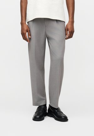 Mann trägt hellgraue, maßgeschneiderte Hosen, schwarze Loafers und ein weißes, strukturiertes Hemd, das über der Taille gekürzt ist, und steht vor einem weißen Hintergrund.