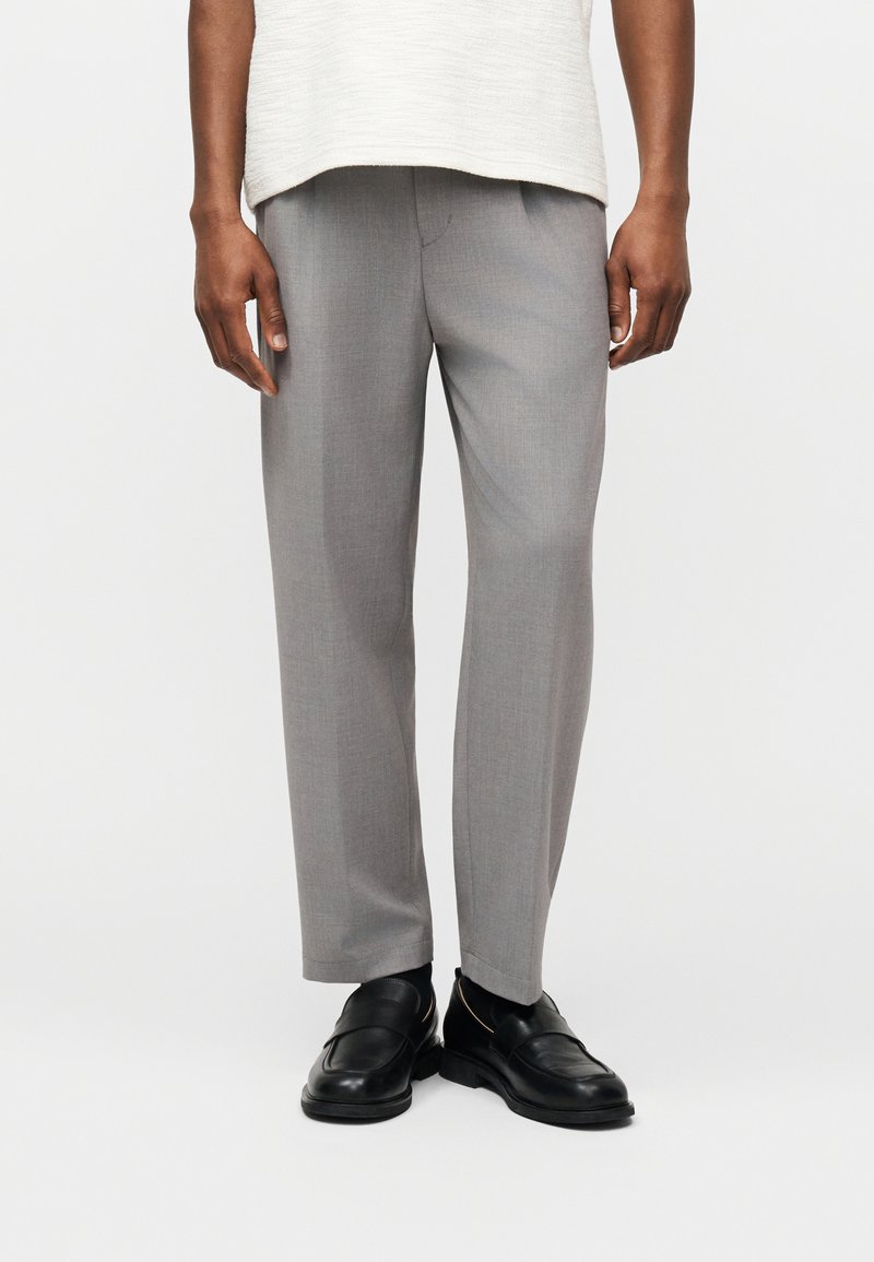 Mann trägt hellgraue, maßgeschneiderte Hosen, schwarze Loafers und ein weißes, strukturiertes Hemd, das über der Taille gekürzt ist, und steht vor einem weißen Hintergrund.