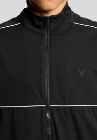 Μαύρο μπουφάν με φερμουάρ και λευκή επένδυση στο στήθος και στους ώμους, με μικρό λογότυπο αετού στο αριστερό μέρος του στήθους.