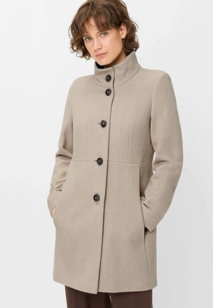 Woman wearing a beige high-collar coat with large black buttons, hands in pockets, standing against a plain light background.