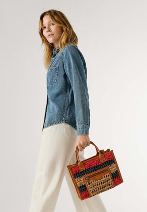 Joven mujer con camisa de mezclilla azul y pantalones blancos sosteniendo a su lado un bolso tejido multicolor con asas de cuero.