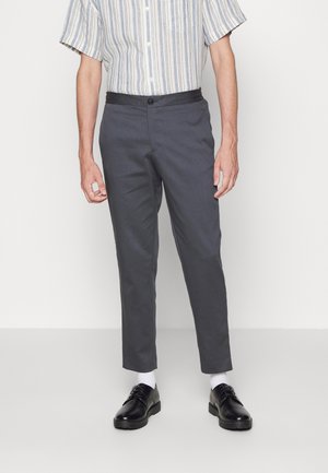 Grijze taps toelopende broek met een gladde textuur, gecombineerd met zwarte schoenen en witte sokken. Gestreept shirt met korte mouwen, knopen en een zak.