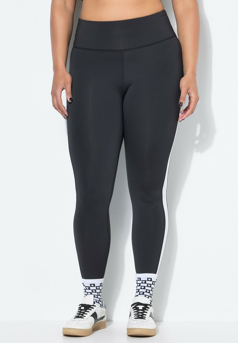 Leggings neri con vita alta, caratterizzati da strisce laterali bianche. Il materiale è elastico e liscio, indossati con calze a fantasia e sneakers.