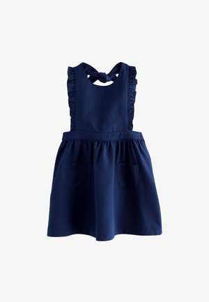Junges Mädchen mit Dutt, das ein dunkelblaues Kleid mit Rüschen, eine weiße Bluse, verspielte Socken und schwarze Schuhe trägt und neben Holzklötzen steht.