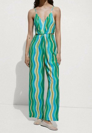 Vrouw die een mouwloze jumpsuit draagt met blauwe, groene en gele verticale golvende strepen, gecombineerd met lichtgekleurde platte schoenen.