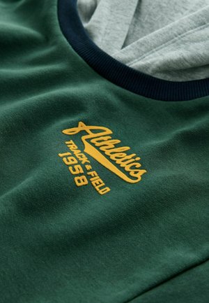 Zaļš krekls ar tumši zilām ribām ap kaklu. Dzeltenā izšūtā tekstā ir rakstīts "Sporta TRACK & FIELD 1958." Mīksta kokvilnas tekstūra.