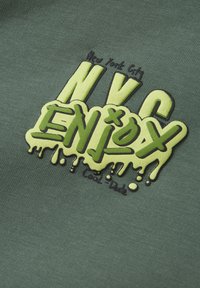 Tela verde con texto impreso en amarillo y negro que dice "NYC ENJ0Y" con un diseño de goteo y acentos, sobre un fondo texturizado.