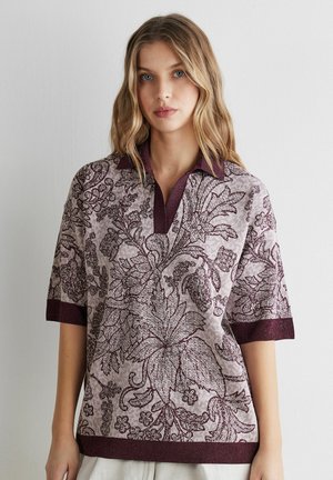 Ung kvinde med blondt hår iført en kortærmet striktop med store blomsterprint i afdæmpede lilla nuancer.