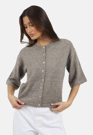 Femme aux longs cheveux foncés portant un cardigan gris à manches courtes boutonné et un pantalon blanc, tenant l'ourlet du cardigan avec les deux mains.