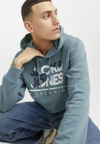 Sudadera con capucha en azul verdoso apagado, con un estampado gráfico "JACK & JONES" en colores en negrita y contrastantes. Tejido suave y corte relajado.