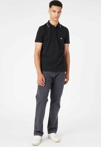 Black polo shirt with white trim, featuring a three-button placket and a small logo on the chest, paired with dark jeans and white sneakers.
