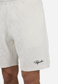 Shorts blancs avec un motif en diamant texturé, taille élastique et logo "Pegador" noir sur l'ourlet gauche.