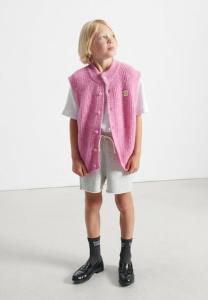 Rozā adīta bezdelīga bez piedurknēm ar ribota tekstūra, ar priekšējo pogu aizdari un mazu logo plāksteri. Nēsāta virs baltas krekliņas un pelēkiem šortiem.