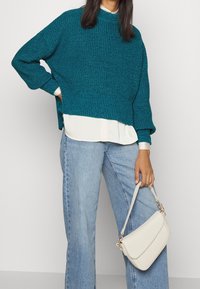 Tyrkysový pletený svetr s vysokým límcem, vrstvený přes bílou košili, doplněný světle modrými džínami se širokými nohavicemi a malou krémovou kabelkou.