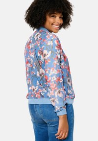 Ziedu bomber jakas gaiši zilos toņos, ar dzīvotspējīgiem rozā un baltiem ziediem, ribotiem aprocēm un apakšmalu, izgatavota no gludas, vieglas auduma.