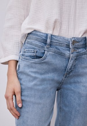 Mujer vistiendo vaqueros de mezclilla azul claro y una blusa blanca texturizada, con la mano apoyada a un lado.