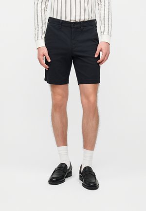 Homme portant un short noir ajusté, des chaussettes blanches, des mocassins noirs et une chemise blanche à manches longues avec des rayures verticales noires.