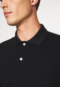 Polo negro con un cuello clásico, dos botones blancos, tela texturizada y un diseño ajustado, mostrado desde la zona del hombro y el cuello.