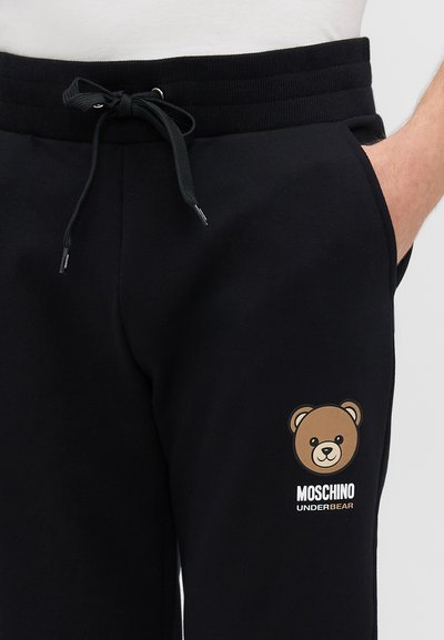Crne trenirke s elastičnom špagom. Sadrže grafiku smešnog medveda i natpis "MOSCHINO UNDERBEAR" u beloj boji na levom nogavici. Mekana tekstura.