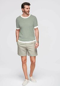 Camiseta de punto a rayas verde y blanca de manga corta, combinada con pantalones cortos gris claro. El modelo lleva zapatillas beiges y un reloj de pulsera.