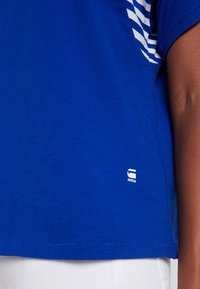 Blue cotton t-shirt featuring white horizontal stripes on the shoulder and a small white logo on the lower left side. Short sleeves, relaxed fit.