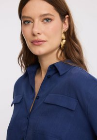 Kobieta o jasnej skórze i niebieskich oczach, ubrana w niebieską koszulę z guzikami oraz dużymi złotymi kolczykami w kształcie liści, patrzy nieznacznie obok kamery.