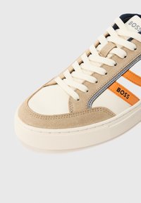 Beige-weißer Sneaker mit cremefarbenen Schnürsenkeln, orange- und grauen Seitenstreifen sowie dem "BOSS"-Logo auf dem orangen Streifen und der Zunge.