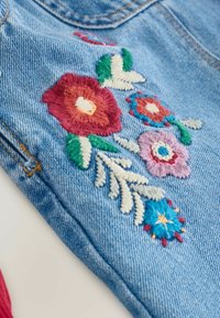 Blauer Denim-Stoff mit buntem, floralem Stickereimuster in Rot-, Pink-, Grün- und Blautönen, verziert mit detaillierten Nähten und Blattelementen.