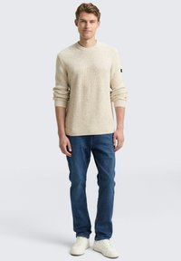 Hellbeige Strickpullover mit gerippter Textur, Rundhalsausschnitt und umgeschlagenen Bündchen, kombiniert mit blauen Jeans und weißen Sneakers.