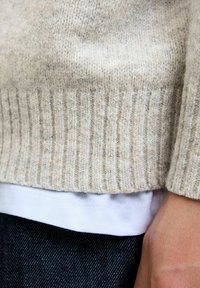 Beige strikket sweater med ribbede kanter, lagd over en hvid skjorte, parret med mørk denim jeans. Blød tekstur og afslappet pasform.