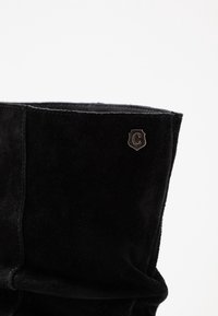 Black suede boot with a soft texture, slightly slouched design, and a silver emblem displaying the letter "C" near the opening.
