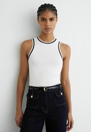 Giovane donna con capelli intrecciati che indossa un top bianco senza maniche con bordi neri e jeans scuri a vita alta con cintura nera, in piedi davanti a un muro bianco.