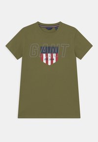 Oliwkowy t-shirt wykonany z bawełny, z napisem "GANT" w szarym kolorze, logo w kształcie tarczy z czerwonymi, białymi i niebieskimi akcentami oraz krótkimi rękawami.
