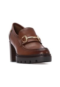 Mocasines de cuero marrón con tacón, punta redondeada, una correa con acento dorado y una suela de plataforma negra gruesa. Presenta un tacón de madera texturizado.