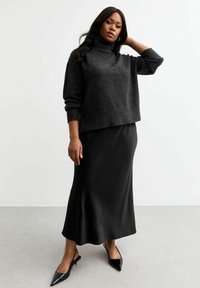 Un pull noir en col roulé avec des manches amples, associé à une longue jupe satin noire et des talons aiguilles noirs, sur un fond clair.