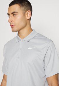 Mlad moški s kratkimi temnimi lasmi, oblečen v svetlo sivo polo majico Nike, gleda levo proti enotni ozadju.
