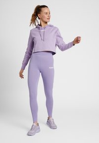 Vrouw staat in een lichtpaarse cropped hoodie, bijpassende high-waist legging en sneakers, kijkt naar rechts tegen een witte achtergrond.
